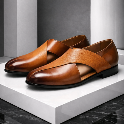 Tan leather men's slip-on loafers with cross strap detail and black sole displayed on white platform