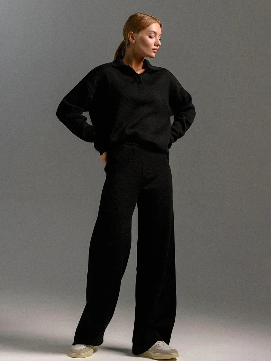 Woman wearing black collared knit sweatshirt and wide-leg pants matching set with white sneakers on gray background.