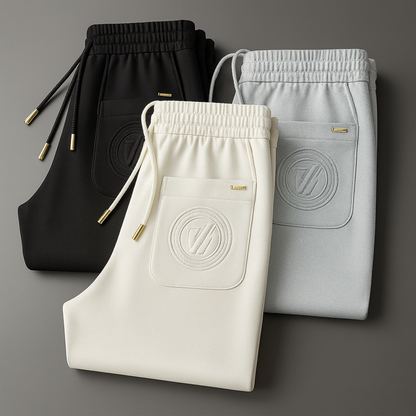 Three folded men's ice silk drawstring sweatpants in black, cream, and gray with gold hardware and embossed logo pocket.