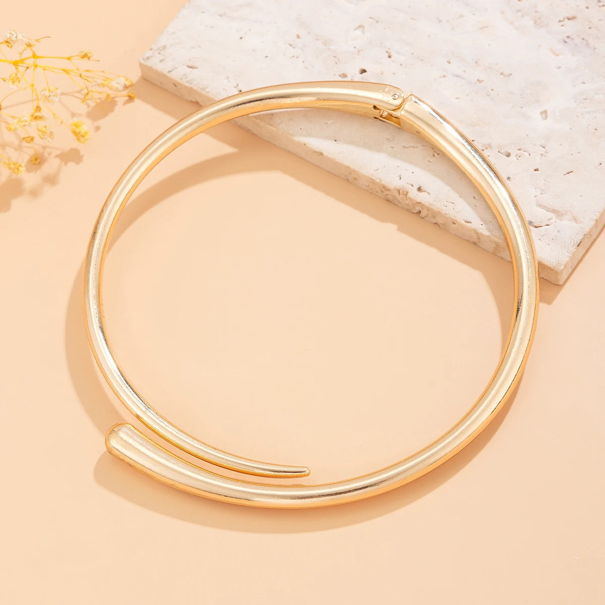 Gold tone open cuff bangle bracelet with modern minimalist design displayed on beige background with flowers