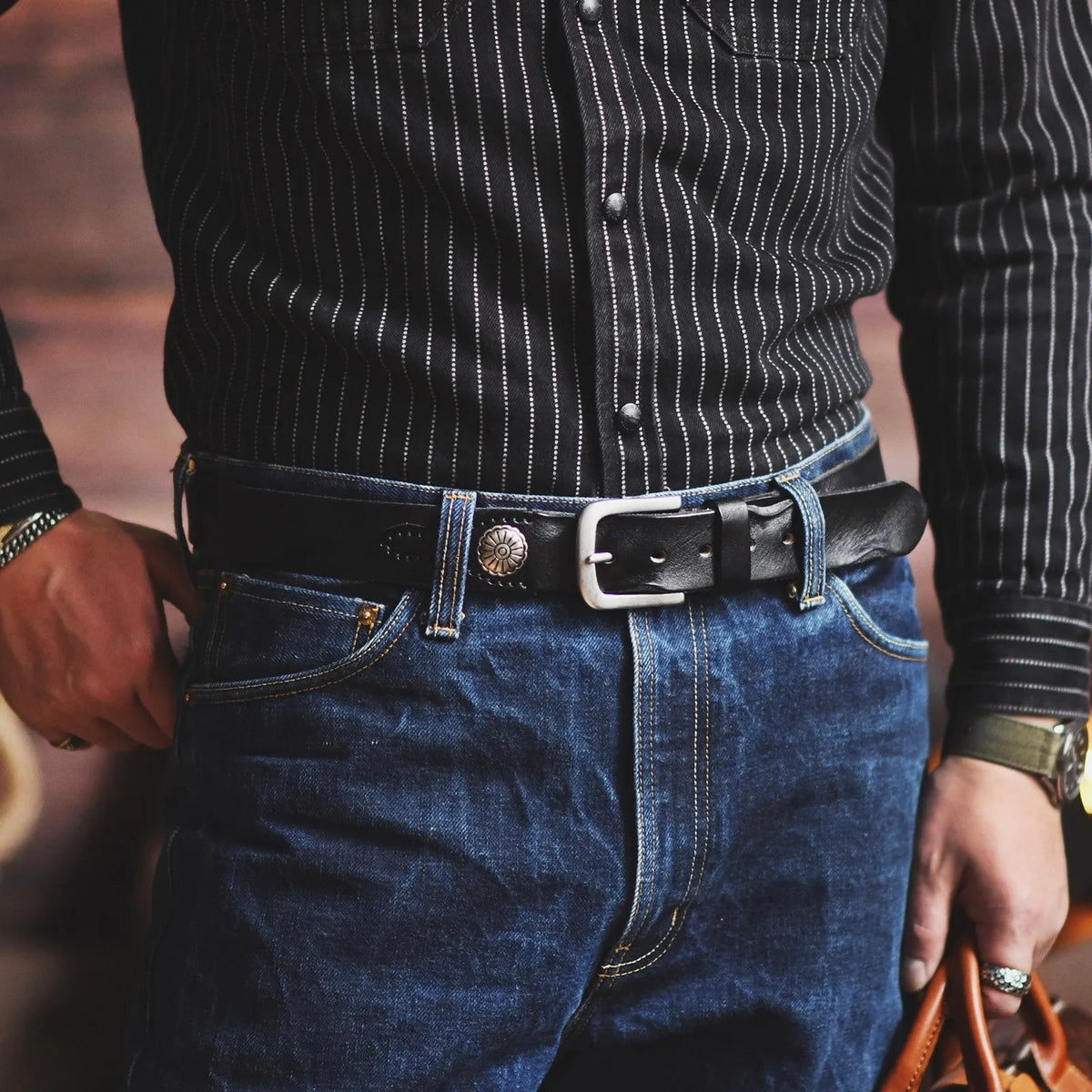 Man wearing black leather belt with silver buckle and decorative metal concho on dark jeans and striped western shirt.