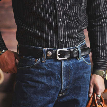 Man wearing black leather belt with silver buckle and decorative metal concho on dark jeans and striped western shirt.