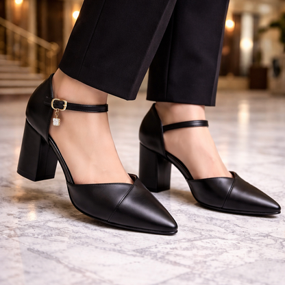 Black leather pointed-toe block heel pumps with ankle strap, gold buckle, and crystal charm detail on marble floor.