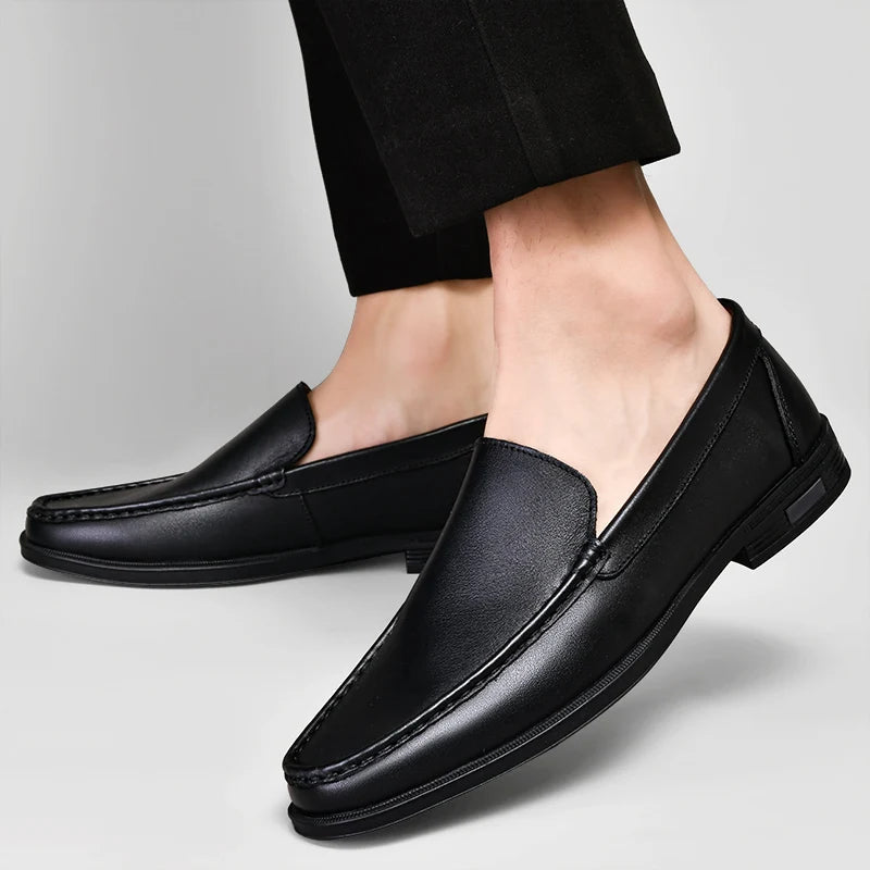 Black men's leather slip-on loafers with contrast stitching, worn with black dress pants on light background.