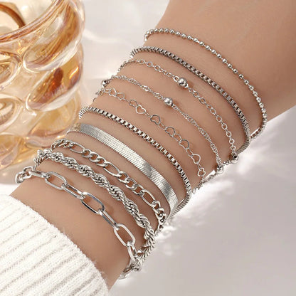 Seven silver chain bracelets stacked on a wrist, featuring varied styles including heart, rope, box, and paperclip chains.