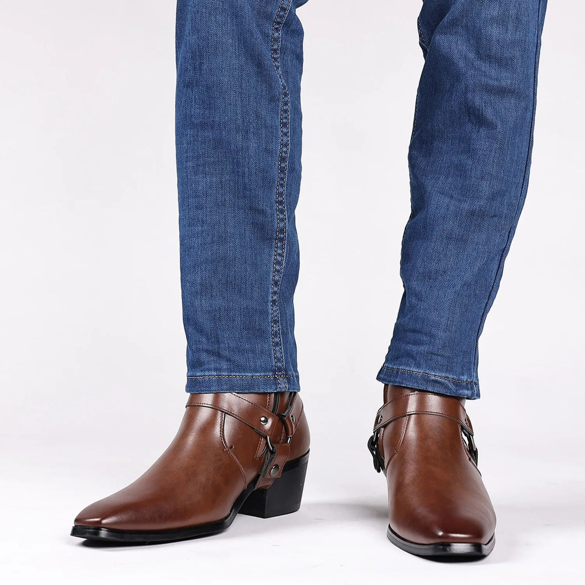 Men's brown leather harness ankle boots with buckle straps and block heel, styled with blue slim-fit jeans.