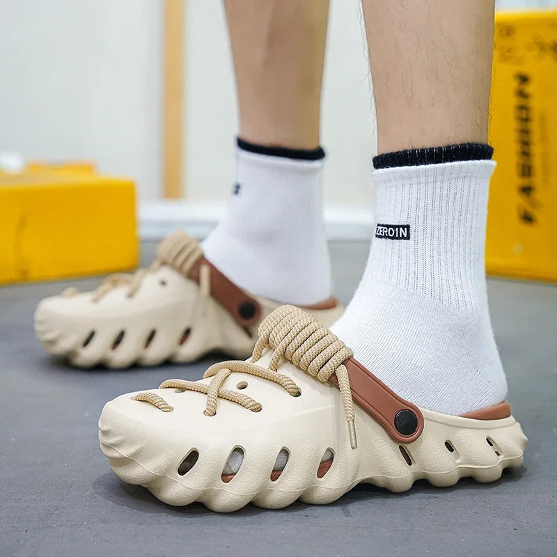 Men's beige cushioned foam lace-up clog shoes with brown strap, worn with white ankle socks on gray surface.
