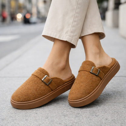 Women's tan plush cotton slip-on mule slippers with silver buckle detail and rubber sole, styled with cream trousers outdoors.