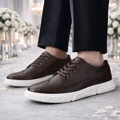 Men's brown leather brogue lace-up sneakers with white rubber sole, worn with black dress pants in elegant floral setting.