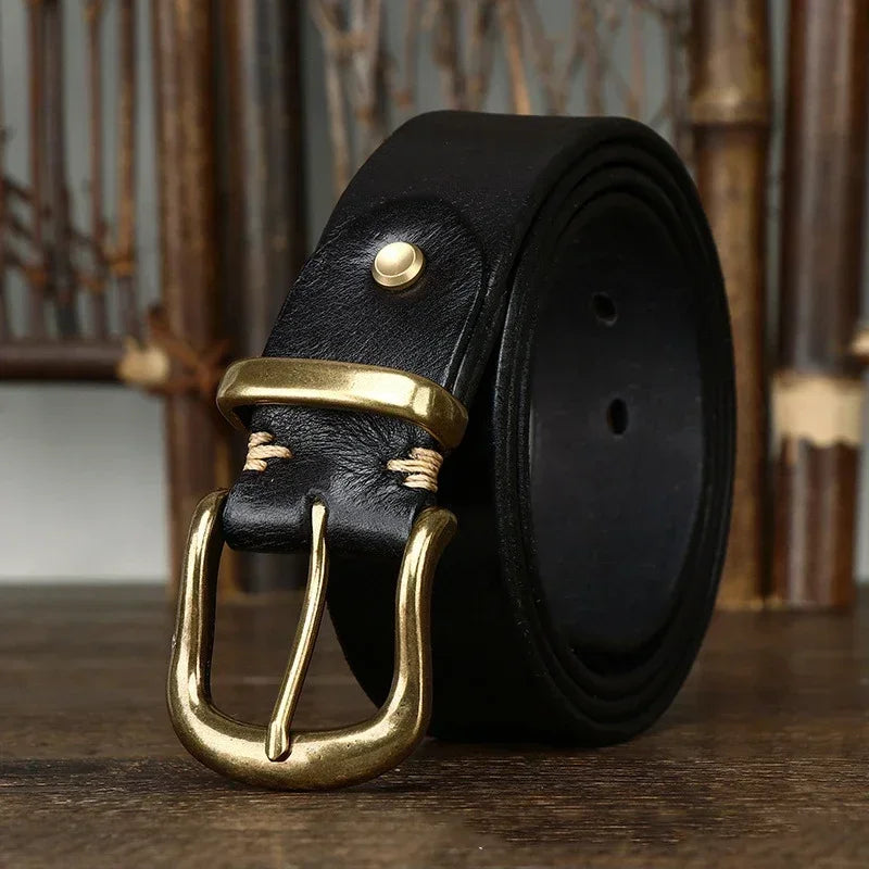 Black genuine leather men's belt coiled with antique brass pin buckle and gold rivet detail on wooden surface