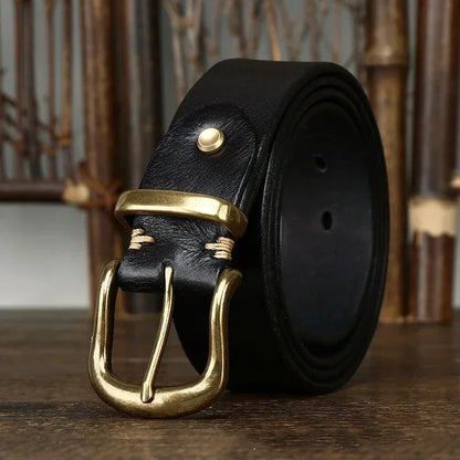 Black genuine leather men's belt coiled with antique brass pin buckle and gold rivet detail on wooden surface