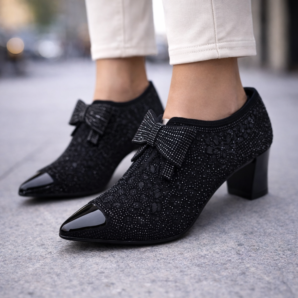 Black rhinestone embellished pointed-toe block heel pumps with bow detail, worn with white cropped pants on pavement.
