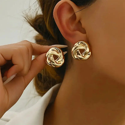 Gold twisted knot stud earrings shown worn on ear and held in hand, featuring interlocked swirl design with snap closure.