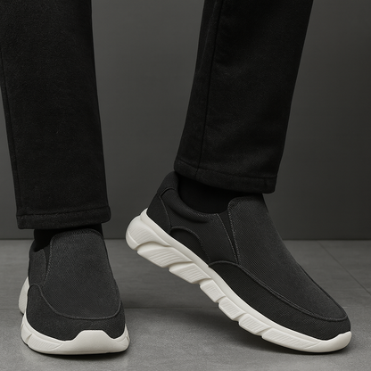 Men's black corduroy slip-on sneakers with white chunky sole, paired with black pants, shown from ankle down.