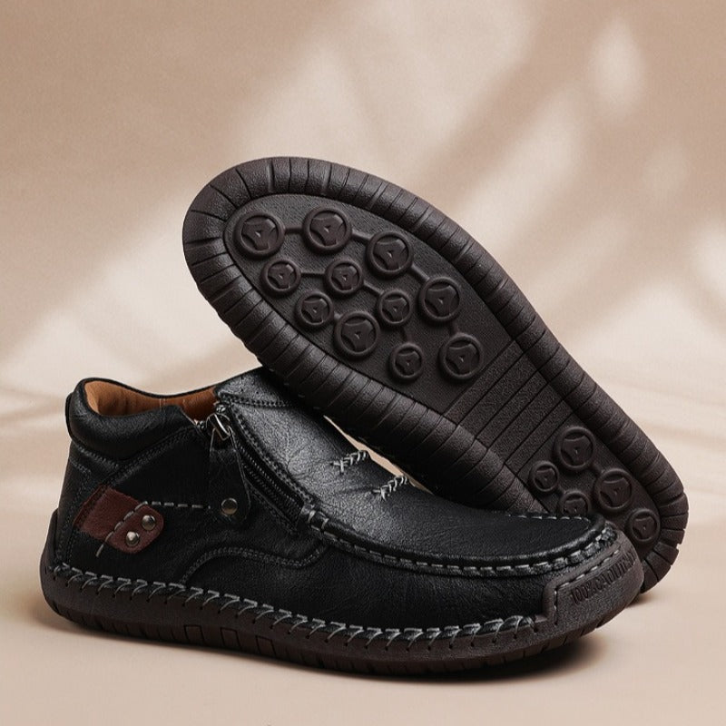 Black leather men's slip-on ankle boots with side zipper, hand-stitching detail, and rubber grip sole displayed on beige background
