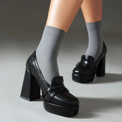Black platform block heel loafers worn with gray crew socks, featuring chunky sole and classic loafer strap detail