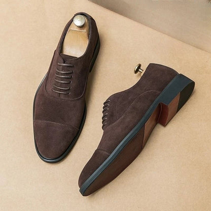 Dark brown suede men's cap-toe Oxford dress shoes with lace-up closure and leather sole, displayed with shoe trees.