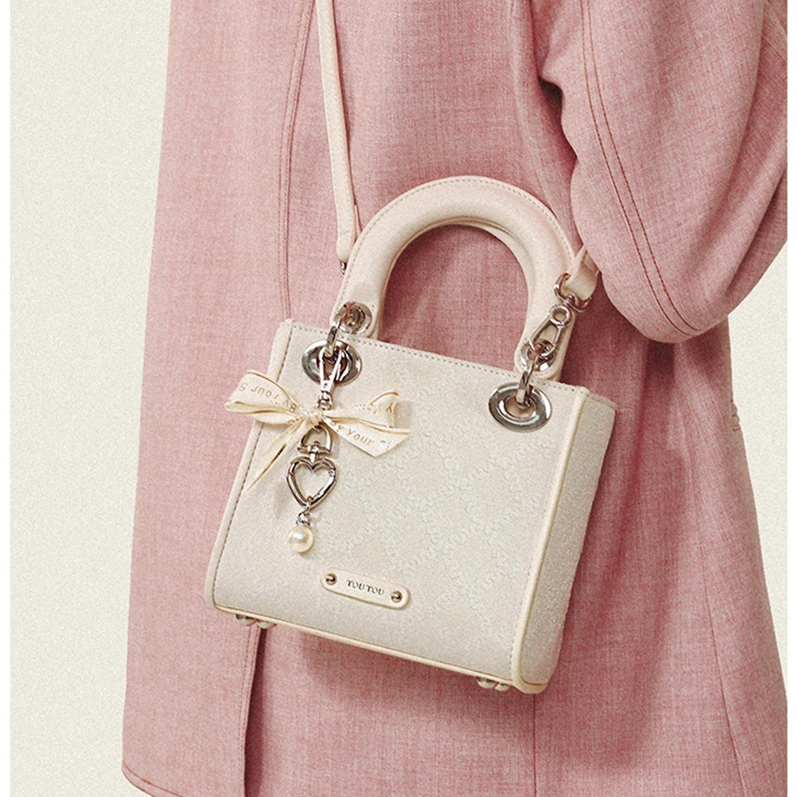 Cream jacquard mini handbag with bow charm, pearl accent, top handles, and crossbody strap against pink blazer background.