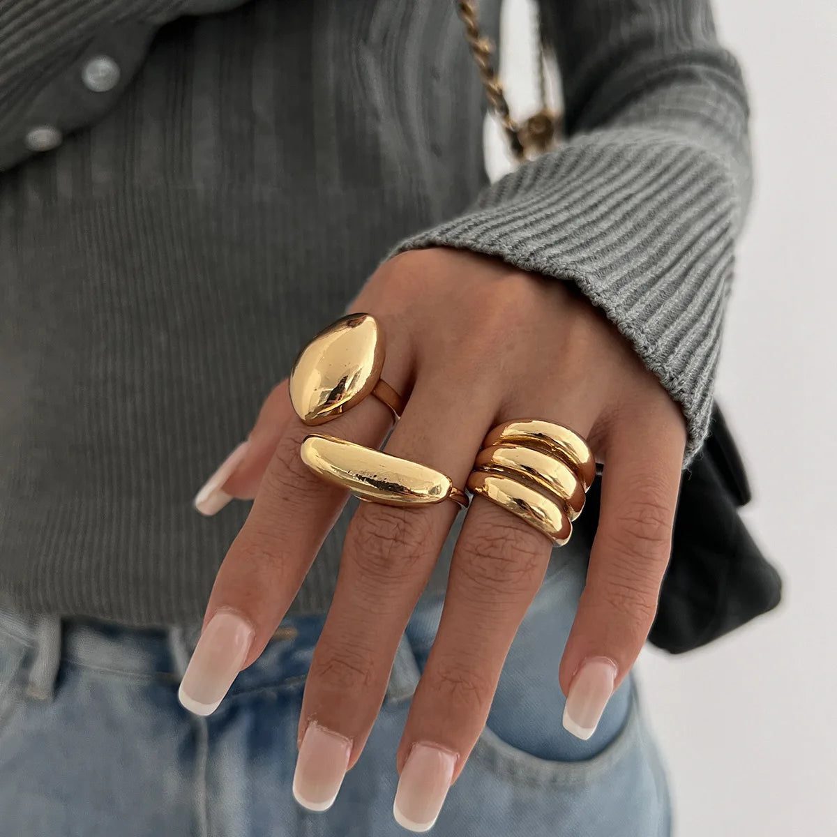 Gold chunky statement ring set on woman's hand featuring dome, oval, and triple-band designs, worn with gray sweater and jeans.