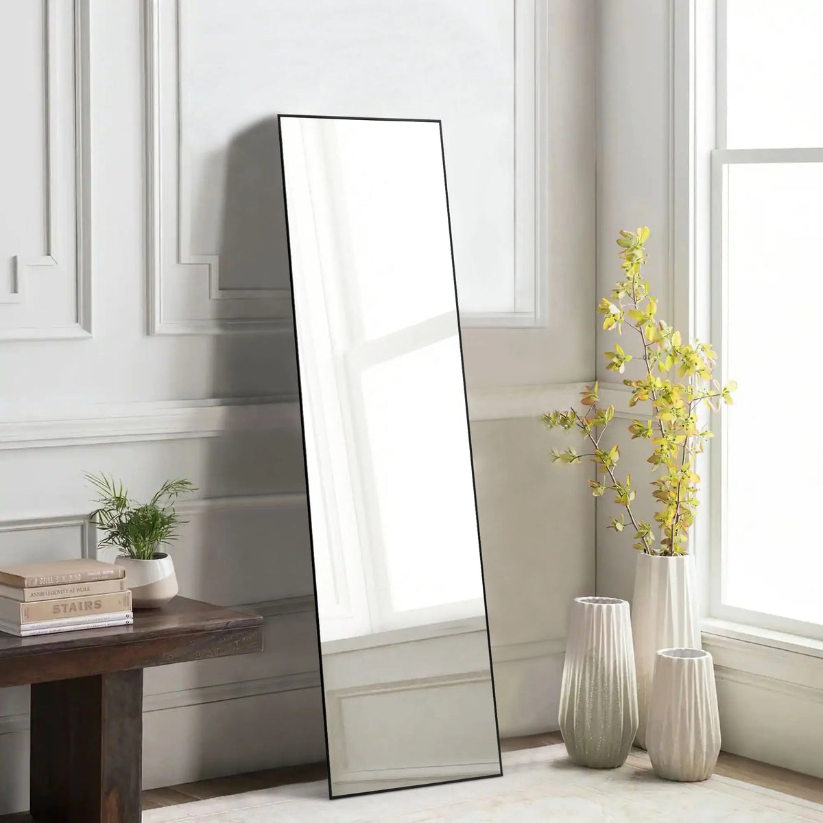 Full length rectangular mirror with black metal frame leaning against white paneled wall beside decorative vases and plants