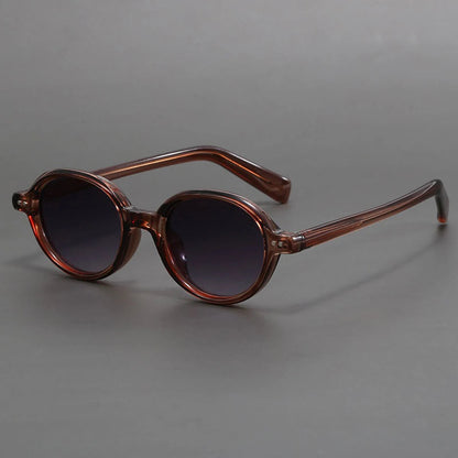 Round sunglasses with transparent brown frames, dark gradient lenses, and slim tortoiseshell temples on gray background