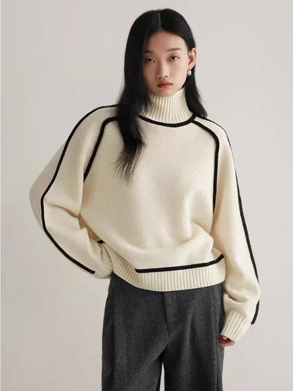Woman wearing cream turtleneck ribbed sweater with black contrast piping, paired with gray wide-leg trousers.