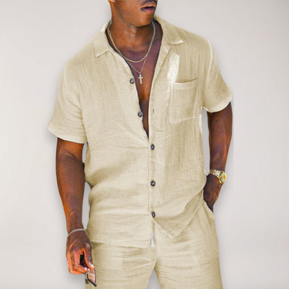 Man wearing beige short-sleeve linen button-up shirt and matching pants, styled with gold jewelry and watch.