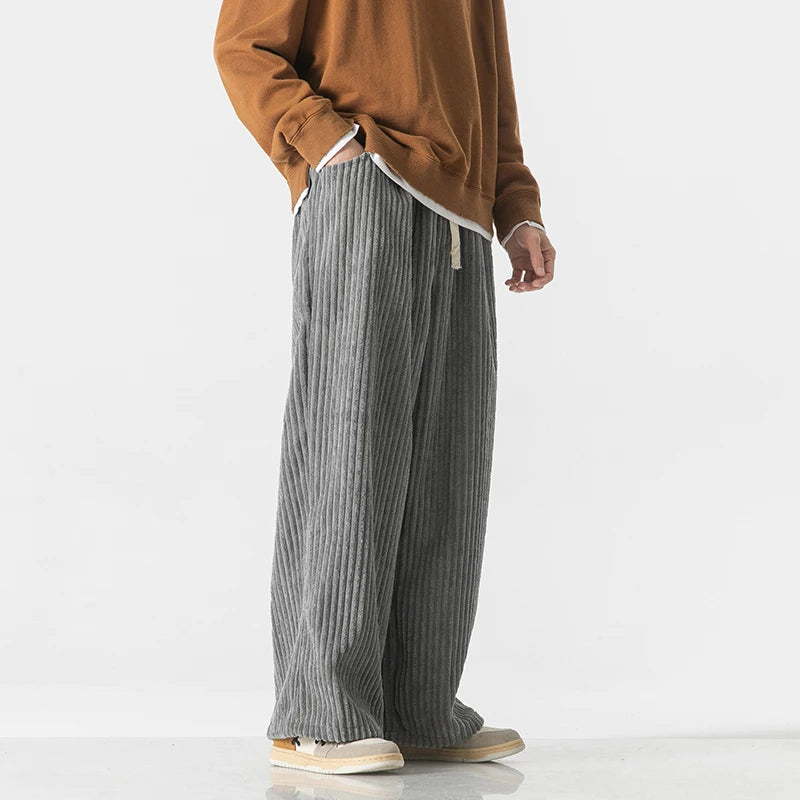 Man wearing gray wide-leg corduroy drawstring pants paired with a brown sweatshirt and white sneakers, side view.