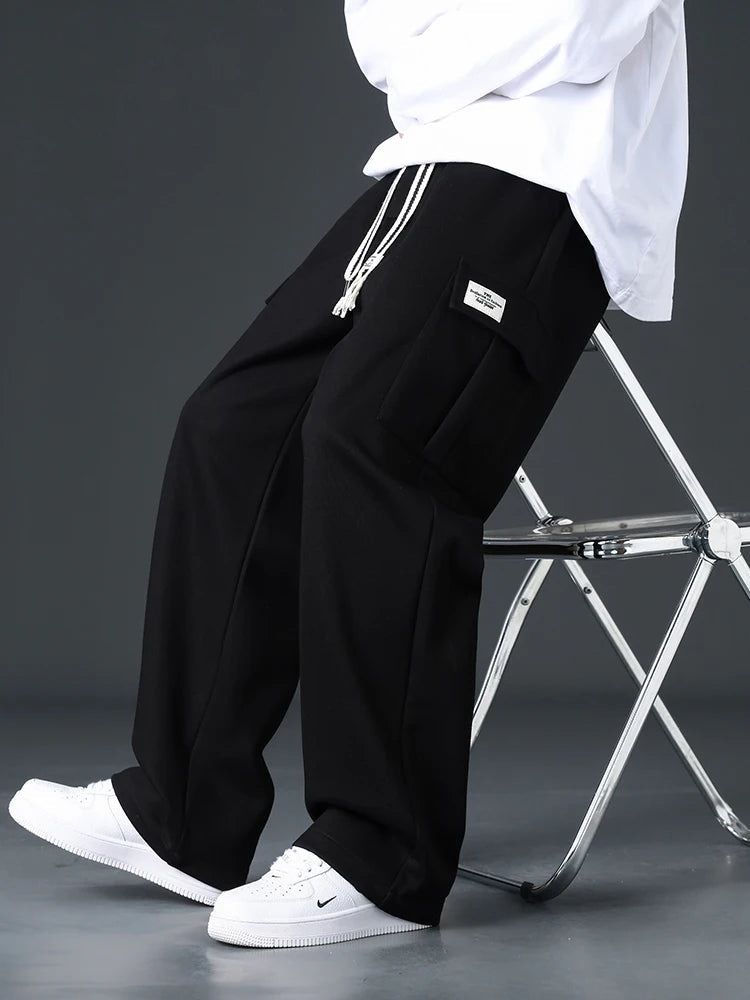Men's black wide-leg sweatpants with drawstring waist and cargo pocket patch, styled with white sneakers and oversized tee.