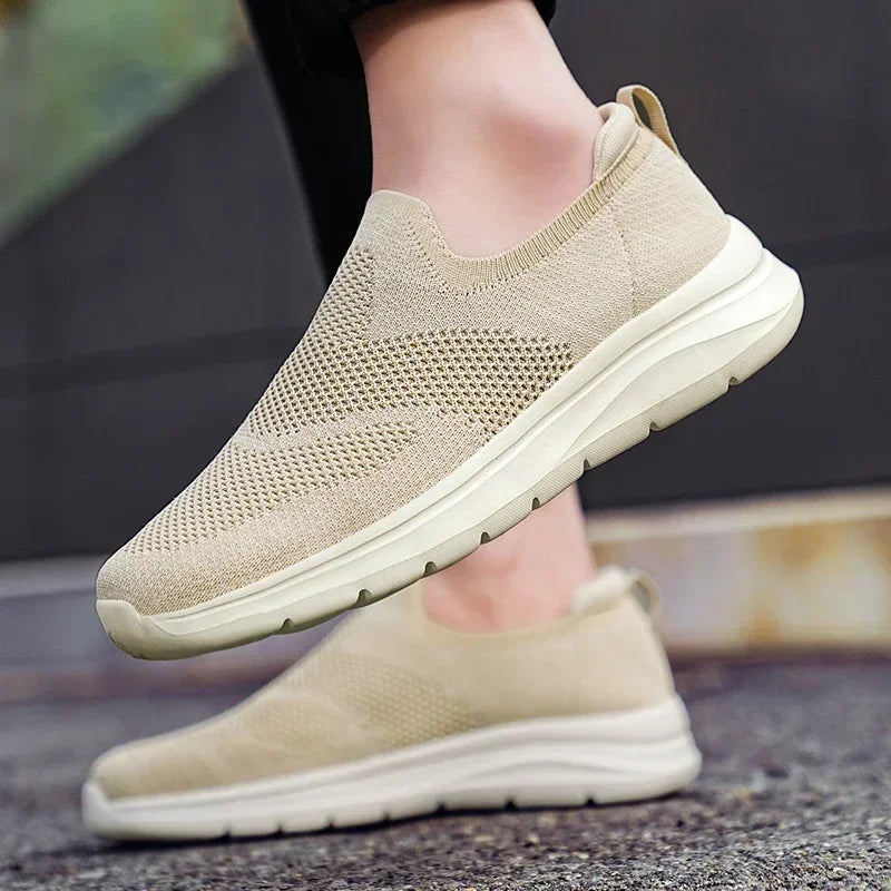 Beige women's slip-on mesh knit sneakers with white sole, shown from side angle on model's feet.