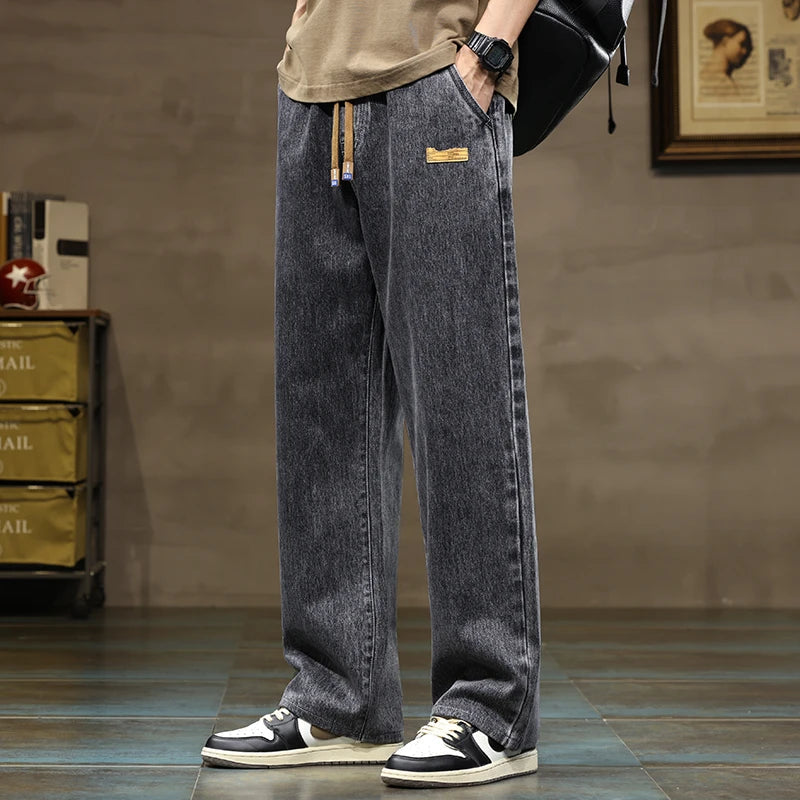 Men's wide-leg drawstring denim jeans in washed gray, paired with a tan tee and black-white sneakers, casual streetwear style.