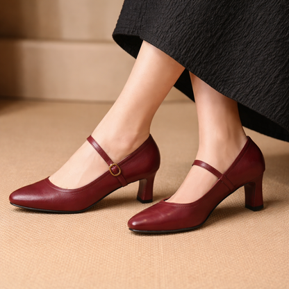 Burgundy leather Mary Jane block heel pumps with ankle strap buckle, worn with a black dress on beige surface.