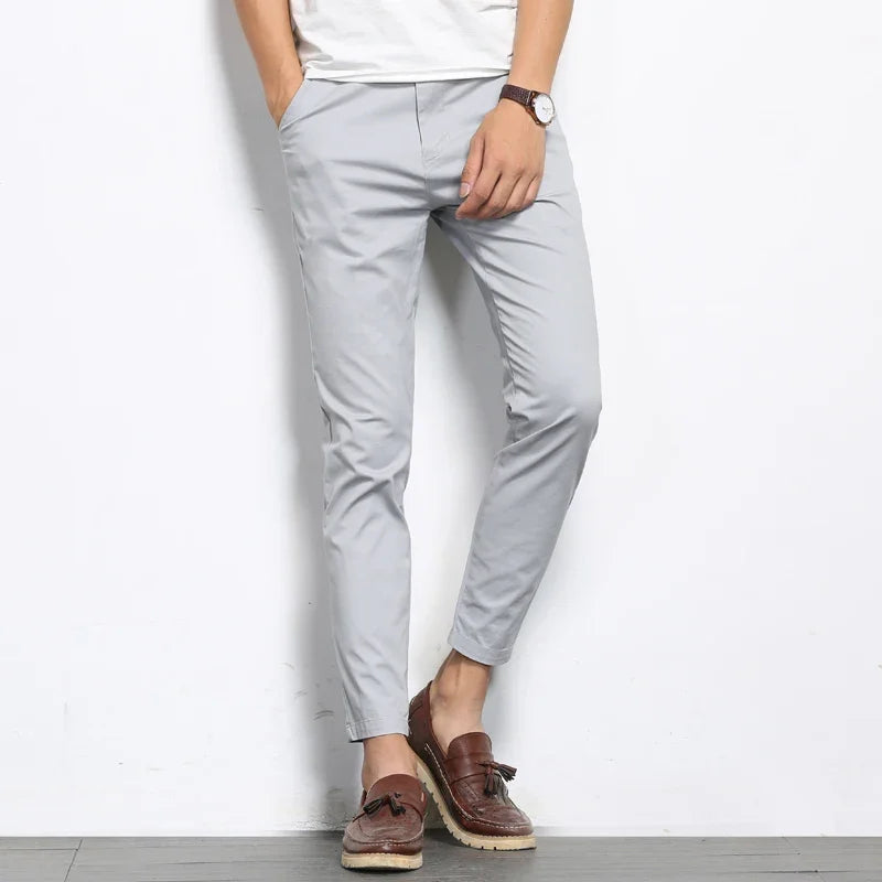Men's light gray slim fit cotton chino pants paired with brown tassel loafers and white tee, cropped ankle length.