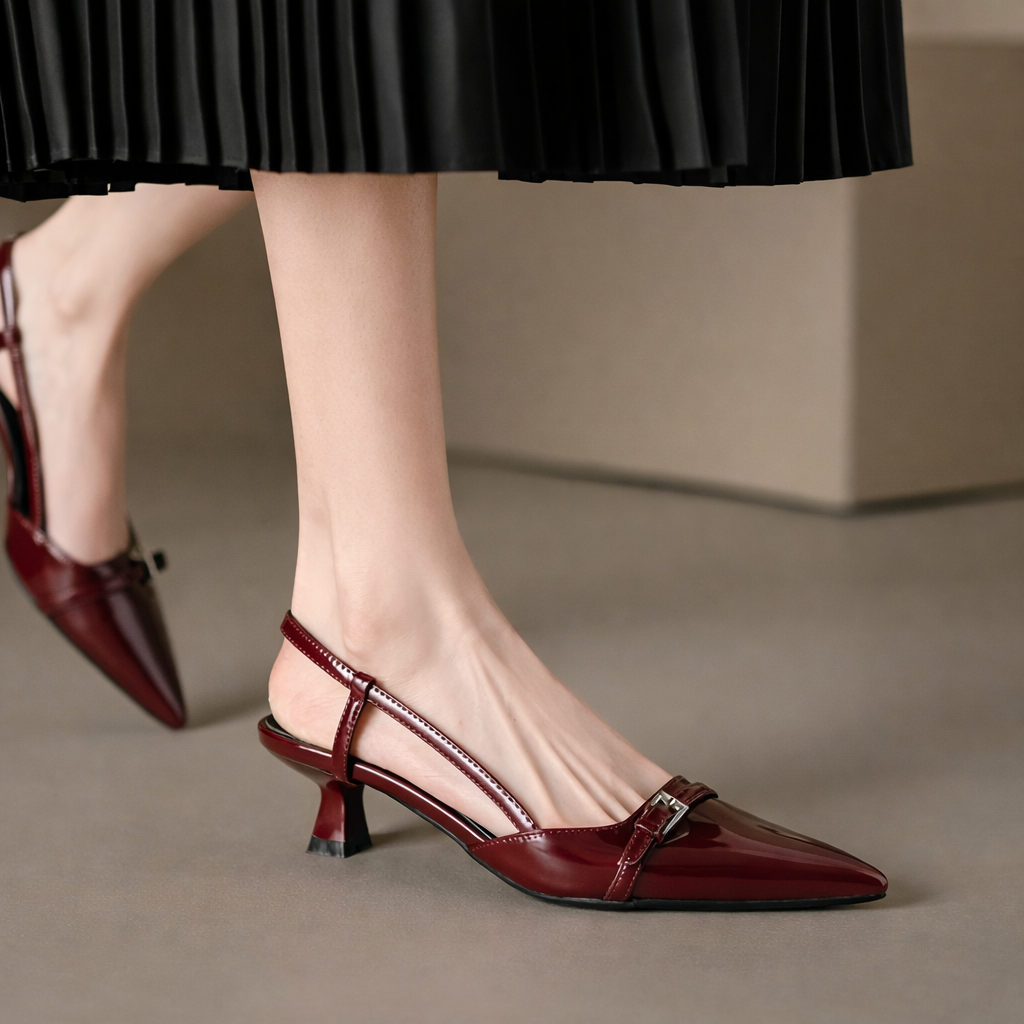 Burgundy patent leather pointed-toe slingback heels with buckle strap detail, worn with a black pleated skirt.