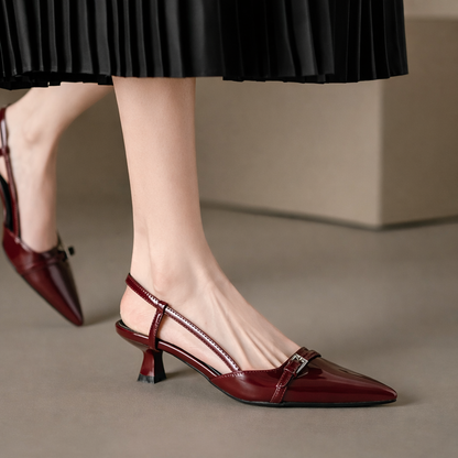 Burgundy patent leather pointed-toe slingback heels with buckle strap detail, worn with a black pleated skirt.