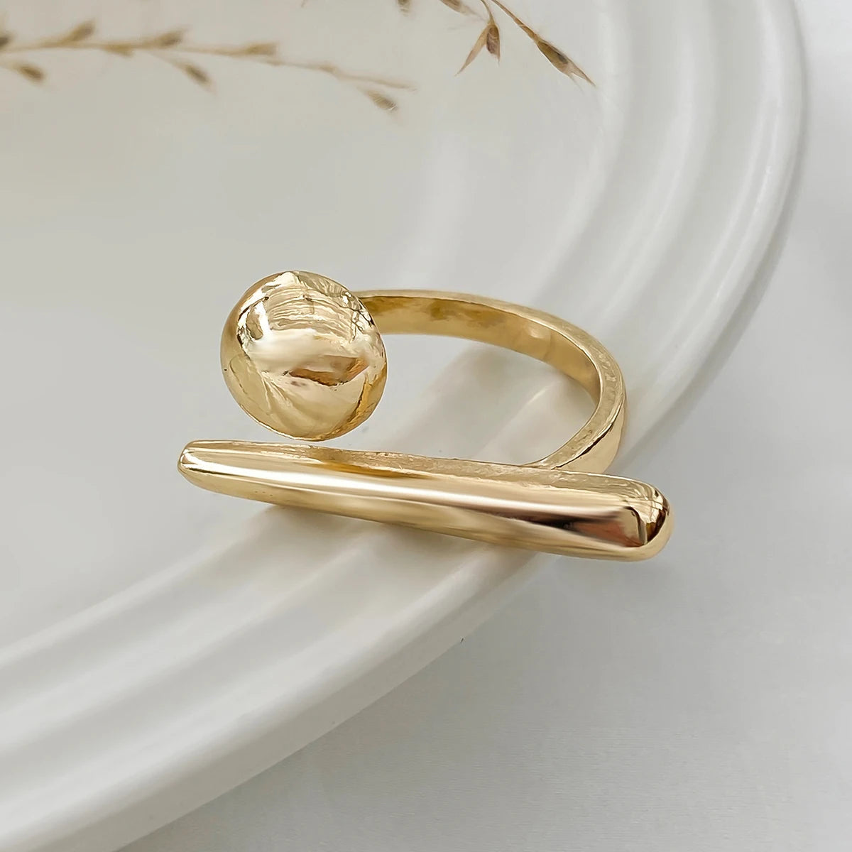 Adjustable gold-tone bar and ball ring for women, modern minimalist open band design on white ceramic plate