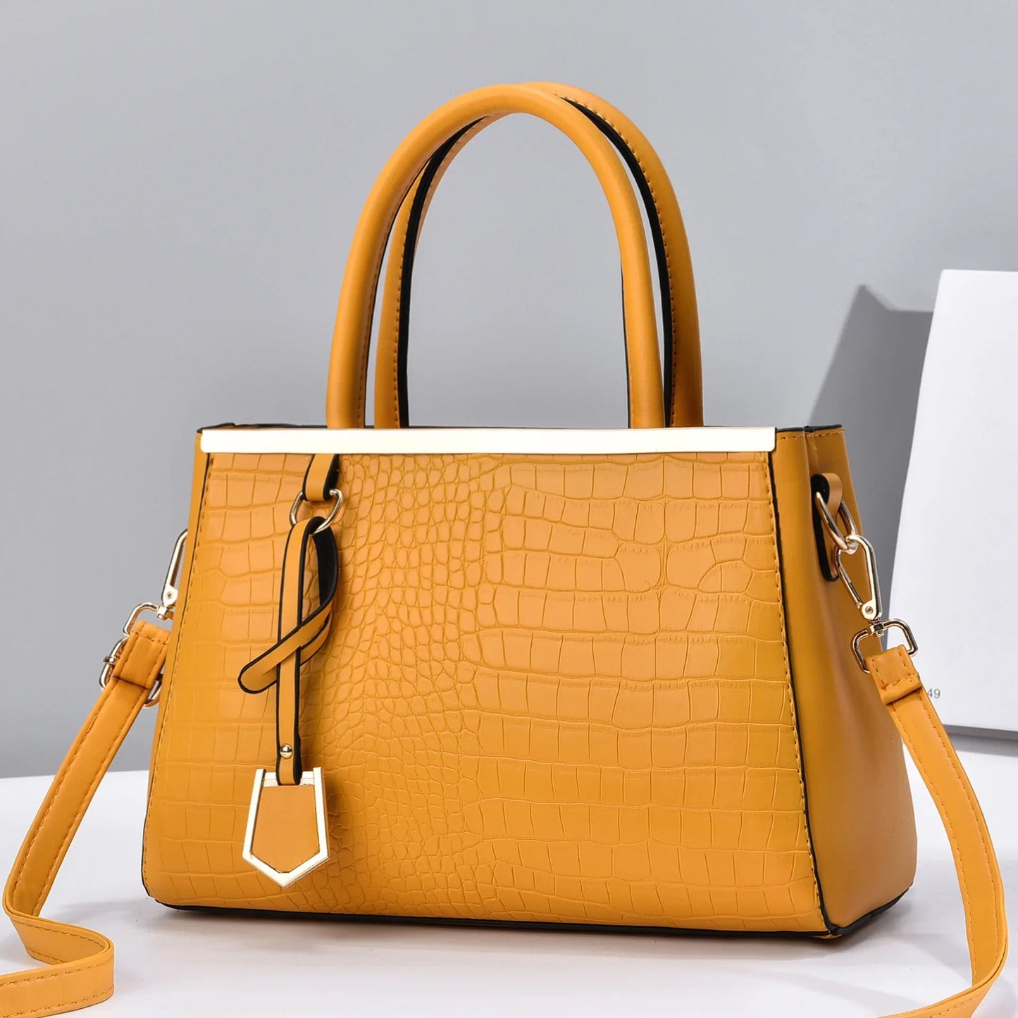 Yellow croc-embossed structured handbag with dual top handles, gold-tone hardware, charm detail, and removable crossbody strap.