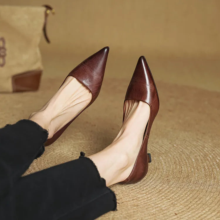 Women's brown leather pointed-toe pumps with kitten heels, styled with black trousers on a woven surface.