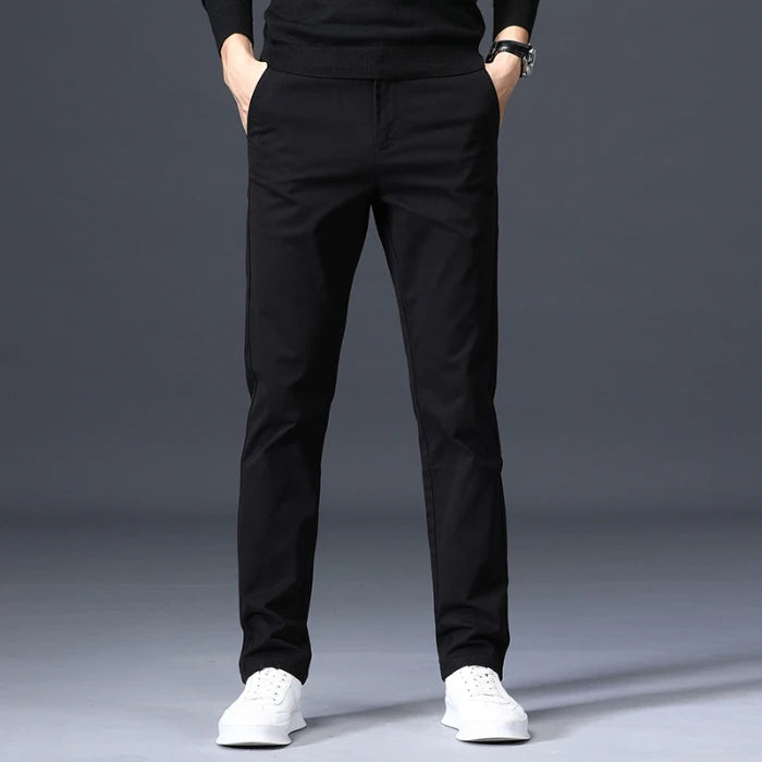Man wearing slim-fit black cotton dress pants with white sneakers and black sweater, hands in pockets, gray background.