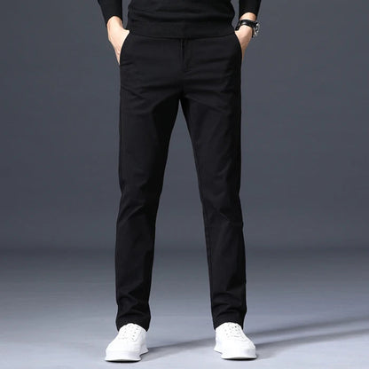 Man wearing slim-fit black cotton dress pants with white sneakers and black sweater, hands in pockets, gray background.
