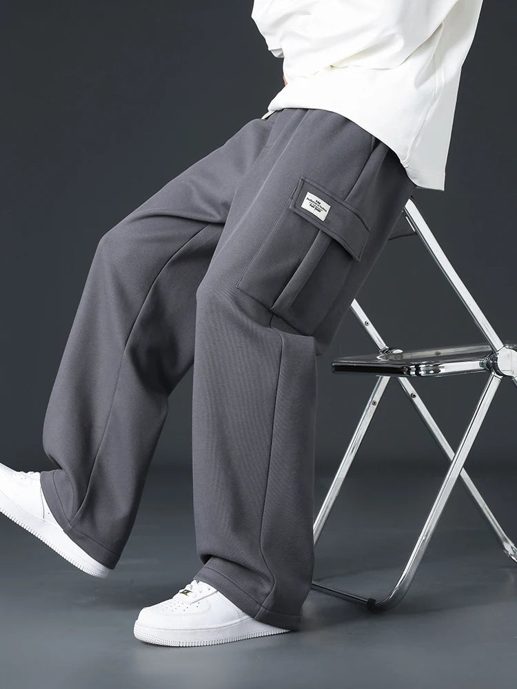 Man sitting on metal stool wearing gray wide-leg cargo sweatpants with side pocket patch label and white sneakers