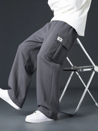 Man sitting on metal stool wearing gray wide-leg cargo sweatpants with side pocket patch label and white sneakers