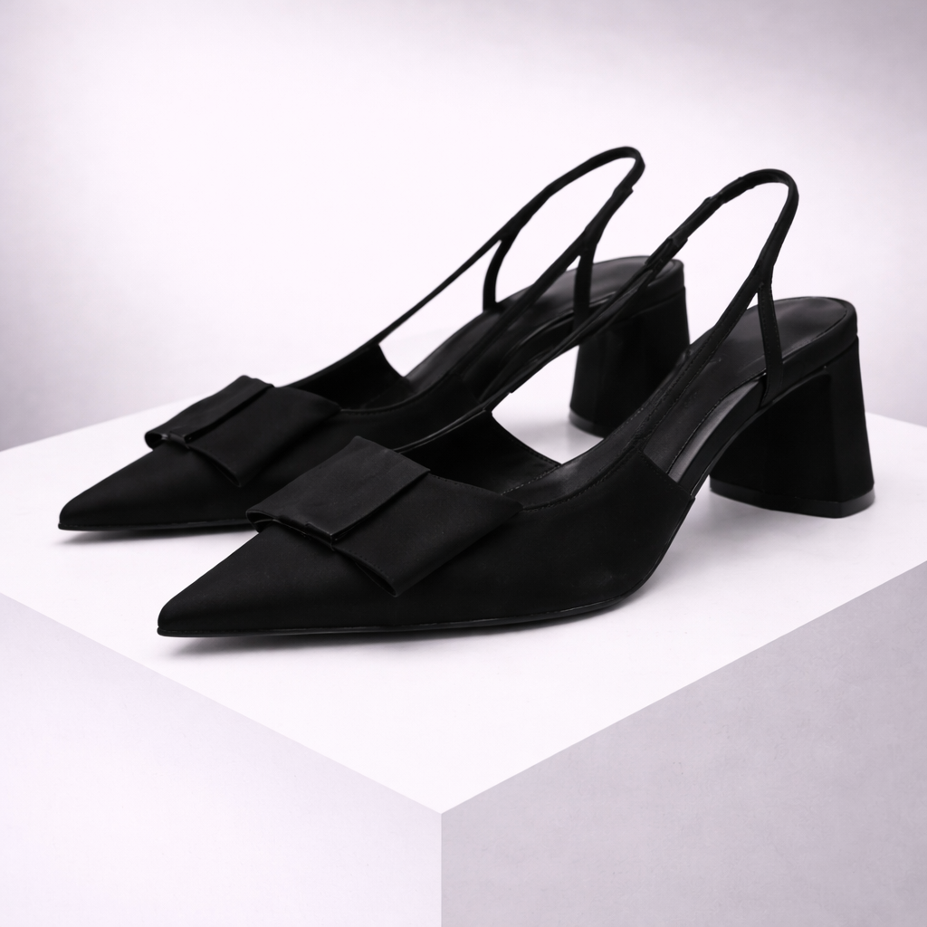 Black satin pointed-toe slingback heels with bow detail and block heel, displayed on white platform