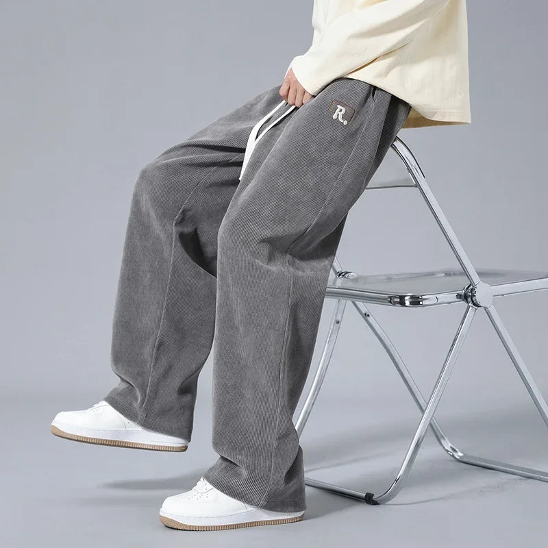 Man sitting on chrome chair wearing gray wide-leg corduroy drawstring sweatpants with R logo patch and white sneakers