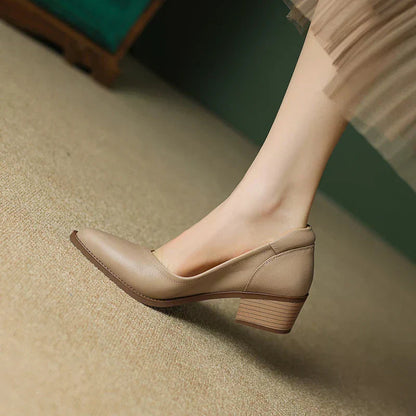 Women's beige leather pointed-toe pumps with a chunky mid-heel, worn with a sheer dress on a neutral floor.