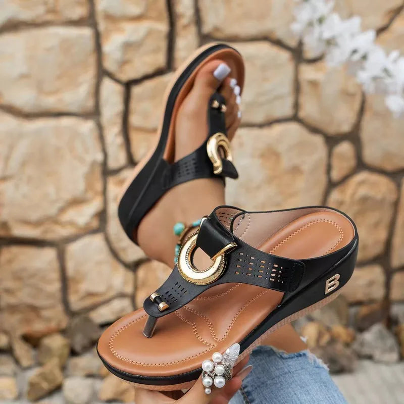 Black wedge T-strap sandals with gold horseshoe buckle detail and tan leather footbed, worn by a woman outdoors.