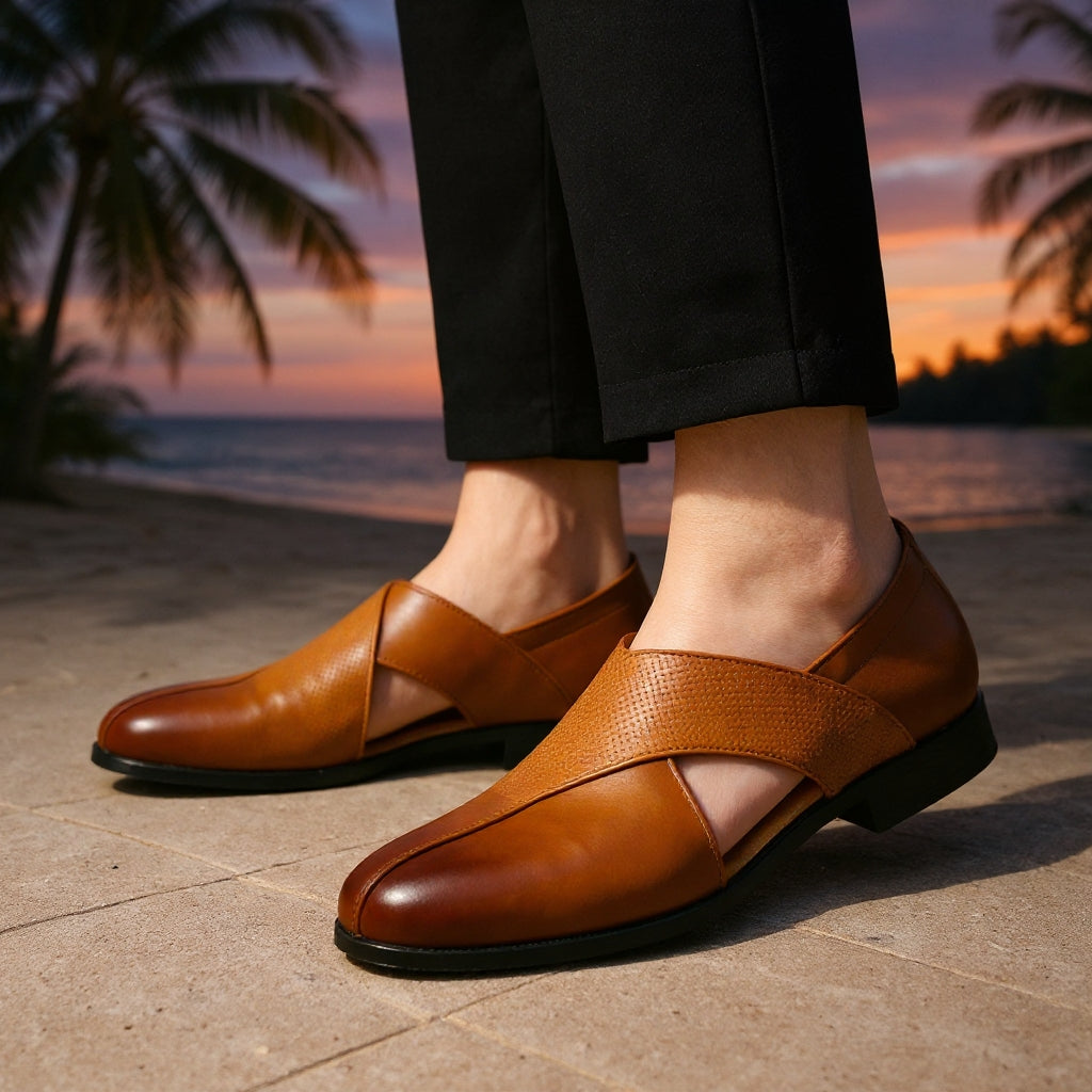 Men's tan leather slip-on loafers with cross strap design, worn with black trousers against a tropical sunset backdrop.