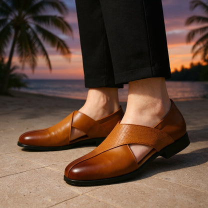 Men's tan leather slip-on loafers with cross strap design, worn with black trousers against a tropical sunset backdrop.