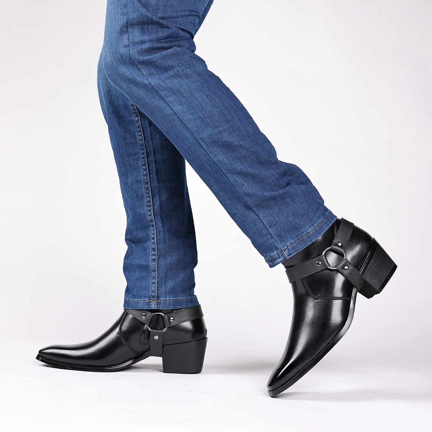 Men's black leather harness ankle boots with block heel and metal ring detail, styled with blue jeans, walking pose.