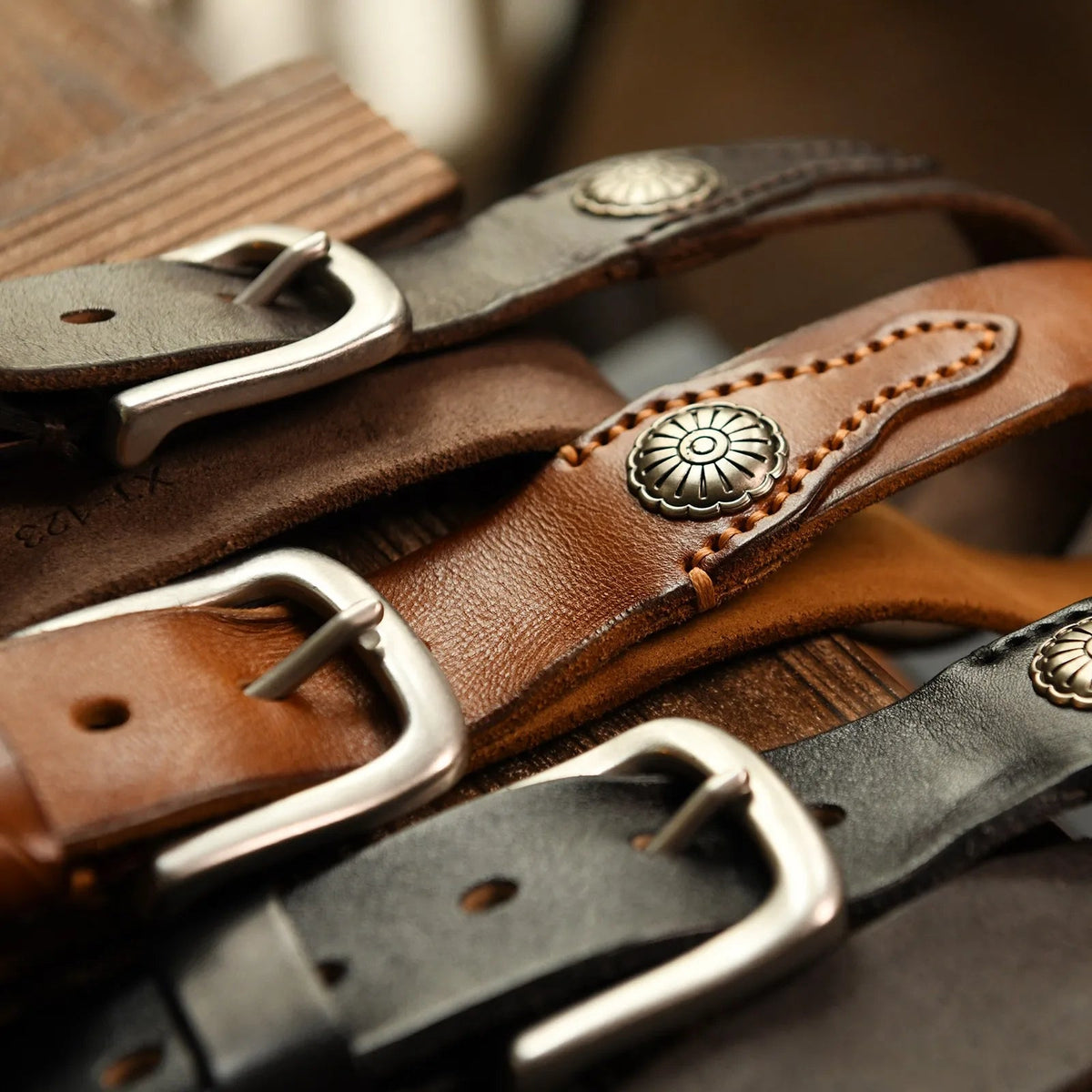 Three stacked leather belts in black, tan, and dark brown with silver buckles and decorative metal concho studs on wood surface.